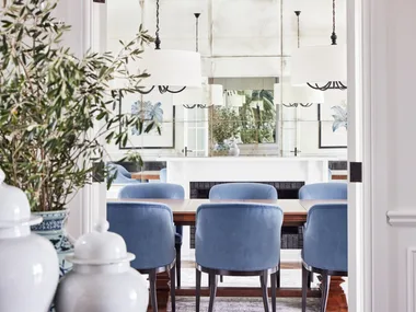 Dining room with blue chairs, white chandeliers, plants, and art on walls, seen through a doorway.
