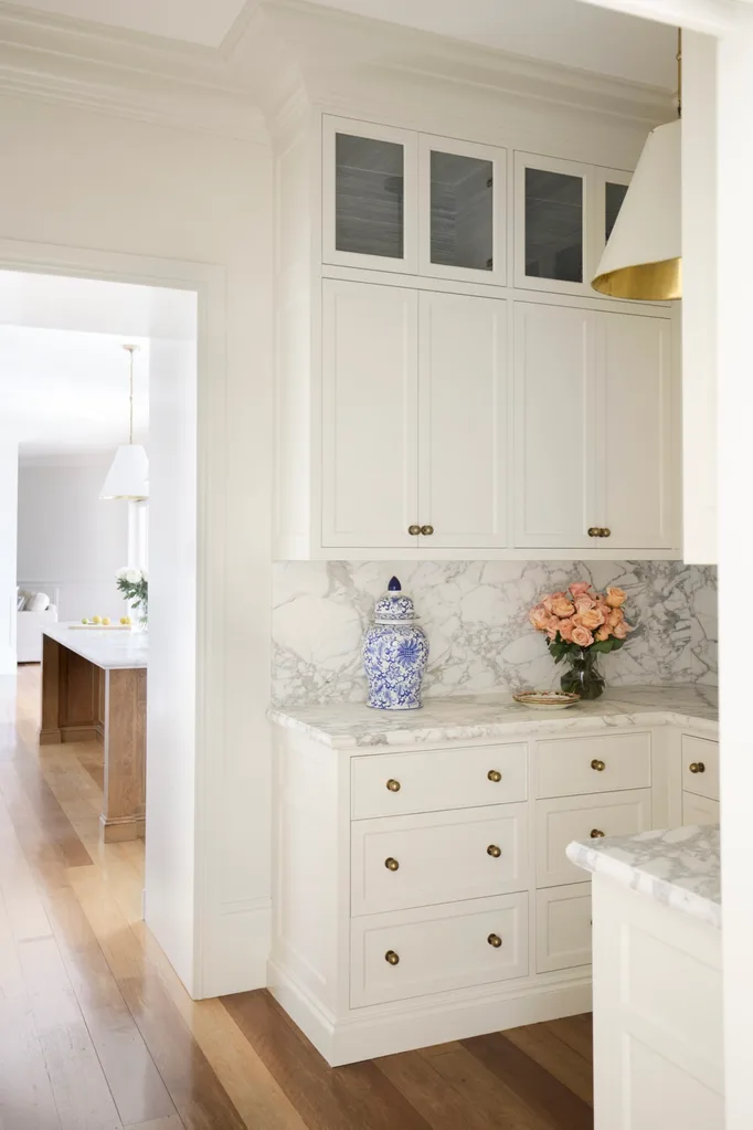 A butler's pantry, with an open doorway leading to the kitchen. Both rooms have timber floorboards, white cabinets, marble countertop with a ginger jar and a vase with pink roses on it.