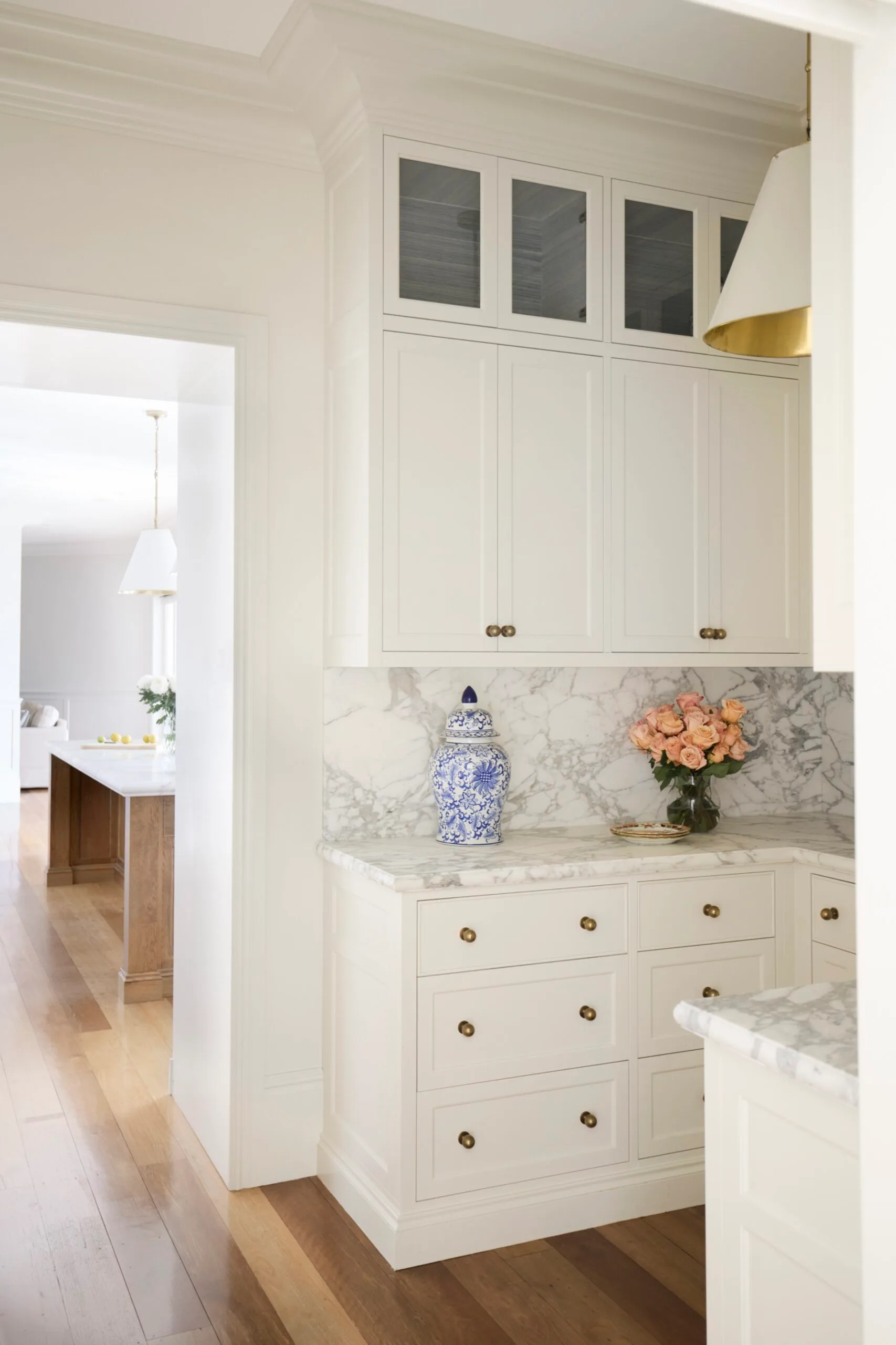 A butler's pantry, with an open doorway leading to the kitchen. Both rooms have timber floorboards, white cabinets, marble countertop with a ginger jar and a vase with pink roses on it.
