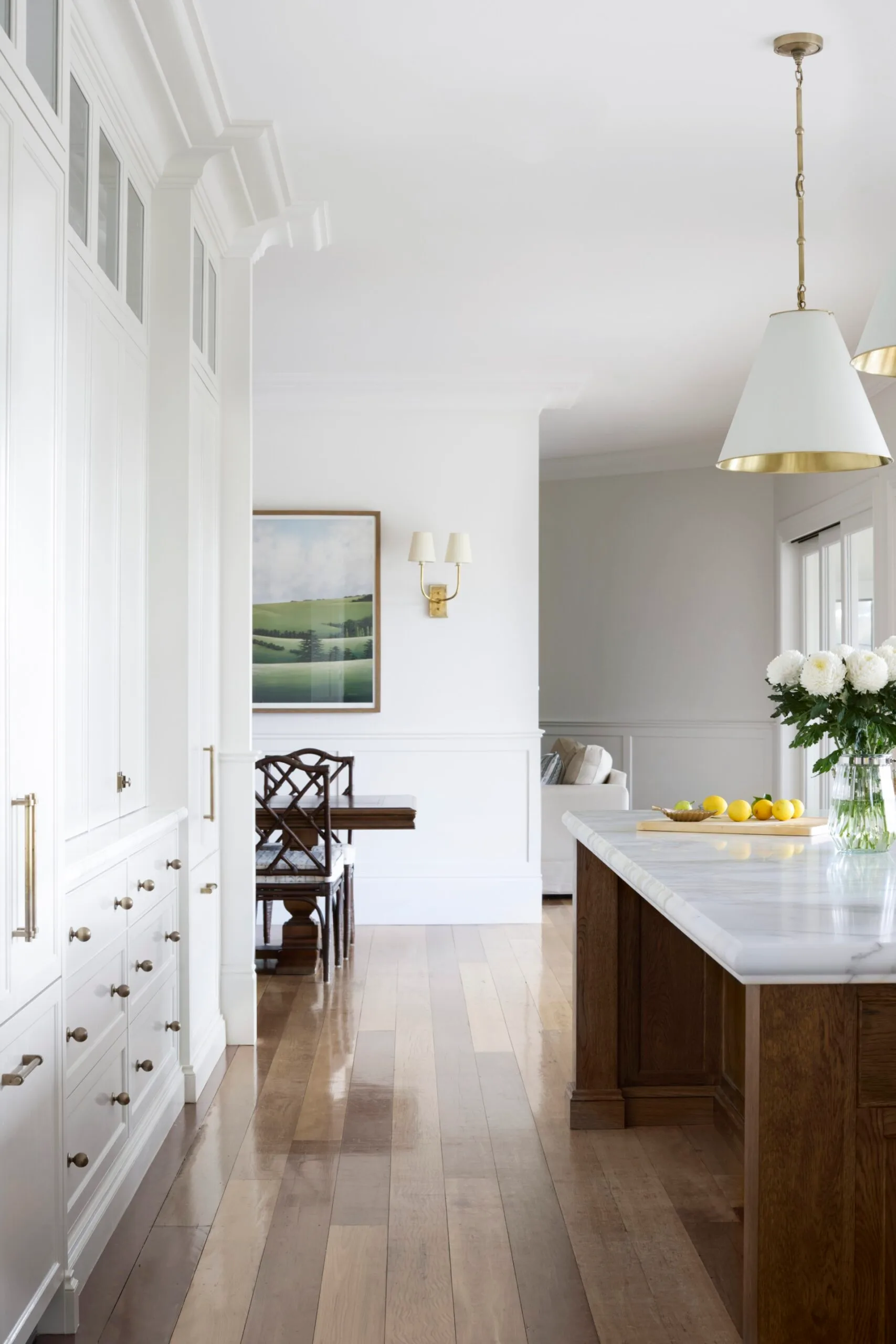Bright kitchen with white cabinets, wooden floors, white pendant lights, dining table in the background. The kitchen island, which has marble countertop, has lemons, and flowers on it. A large artwork depicting the countryside is on the wall near the dining table.