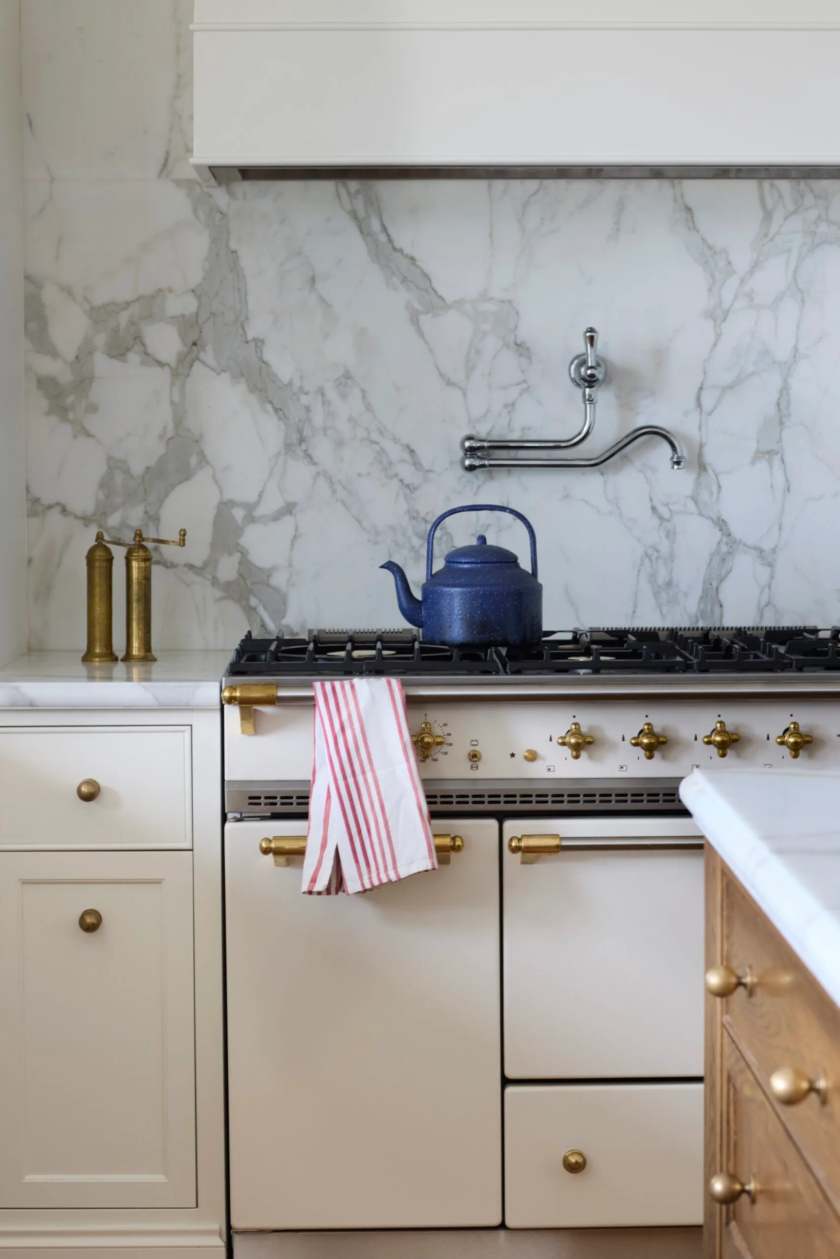 A close up photo of a marble splashback, white shaker joinery with brass knobs, and a Lacanche oven and cooktop with a blue kettle placed on top. A chrome pot filler is on the marble wall.