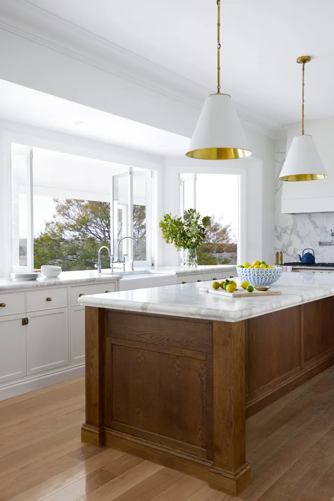 Bright kitchen with white cabinets, wooden floors, white pendant lights above a large kitchen island, which has marble countertop. A bowl of lemons is on it. Windows open out to views of the surrounding countryside.