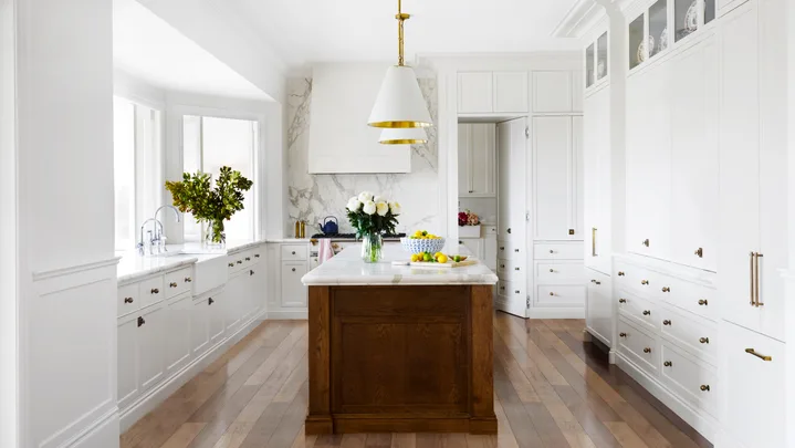 Bright kitchen with white cabinets, wooden floors, white pendant lights above a large kitchen island, which has marble countertop. A bowl of lemons is on it.