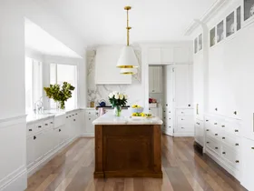 Bright kitchen with white cabinets, wooden floors, white pendant lights above a large kitchen island, which has marble countertop. A bowl of lemons is on it.