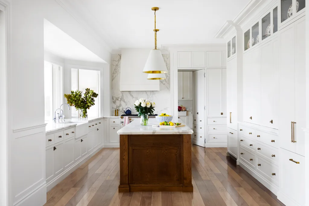 Bright kitchen with white cabinets, wooden floors, white pendant lights above a large kitchen island, which has marble countertop. A bowl of lemons is on it.