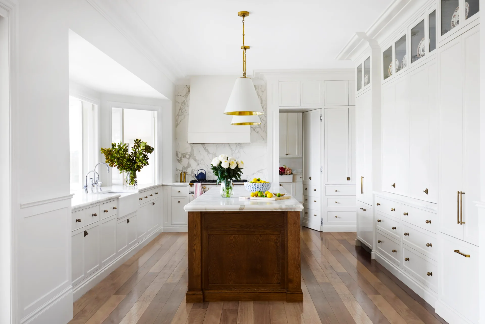 Bright kitchen with white cabinets, wooden floors, white pendant lights above a large kitchen island, which has marble countertop. A bowl of lemons is on it.
