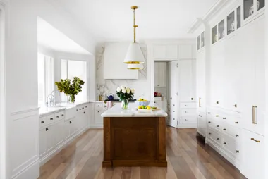 Bright kitchen with white cabinets, wooden floors, white pendant lights above a large kitchen island, which has marble countertop. A bowl of lemons is on it.