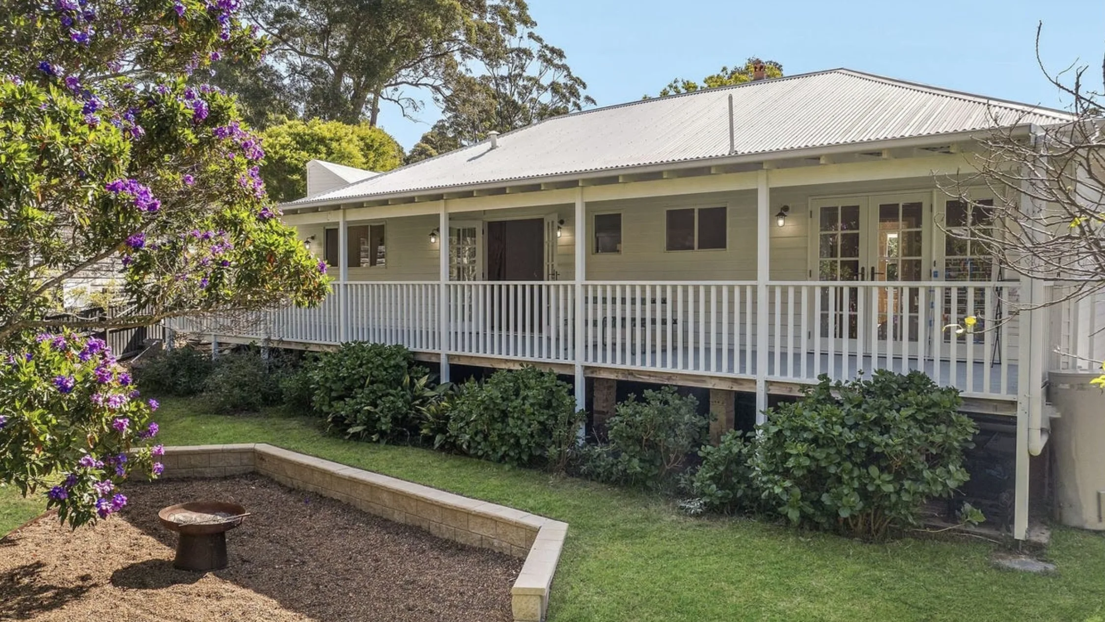 Eleanor Pendleton's White house with a wrap-around porch, surrounded by green shrubs and flowering tree in a garden setting.