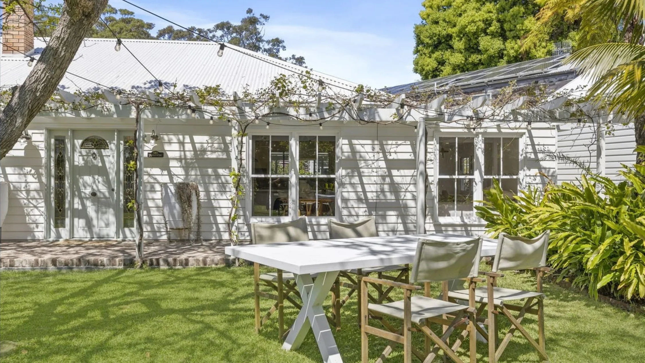 Eleanor Pendleton's White cottage with a white dining table and chairs on a lawn. Solar panels on roof, surrounded by greenery and string lights.
