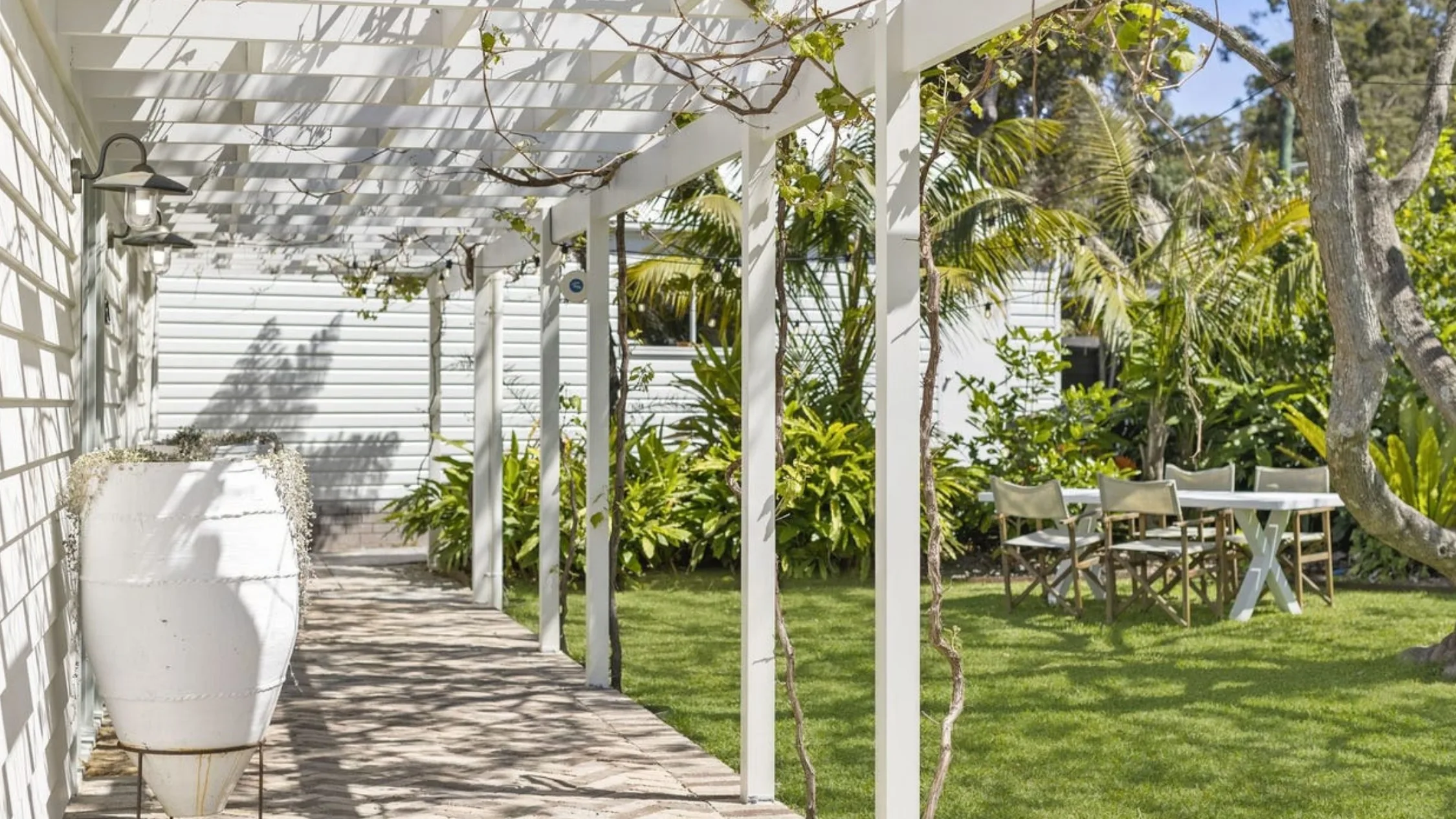 Eleanor Pendleton's Outdoor patio with pergola, white planter, and table with chairs on a neatly trimmed lawn surrounded by lush greenery.