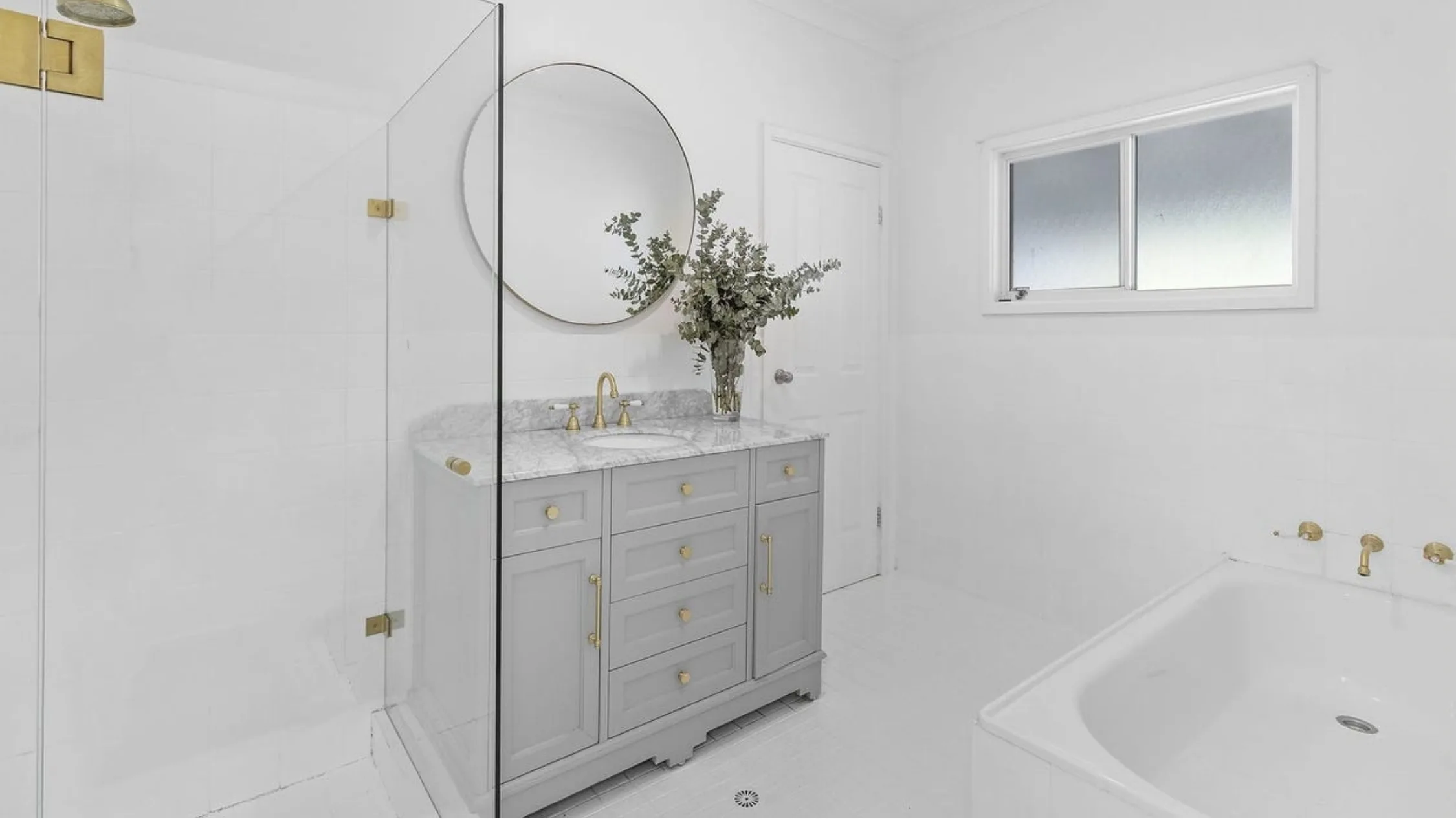 Eleanor Pendleton's Minimalist bathroom with gray vanity, round mirror, glass shower, and white tub; brass fixtures add elegance.