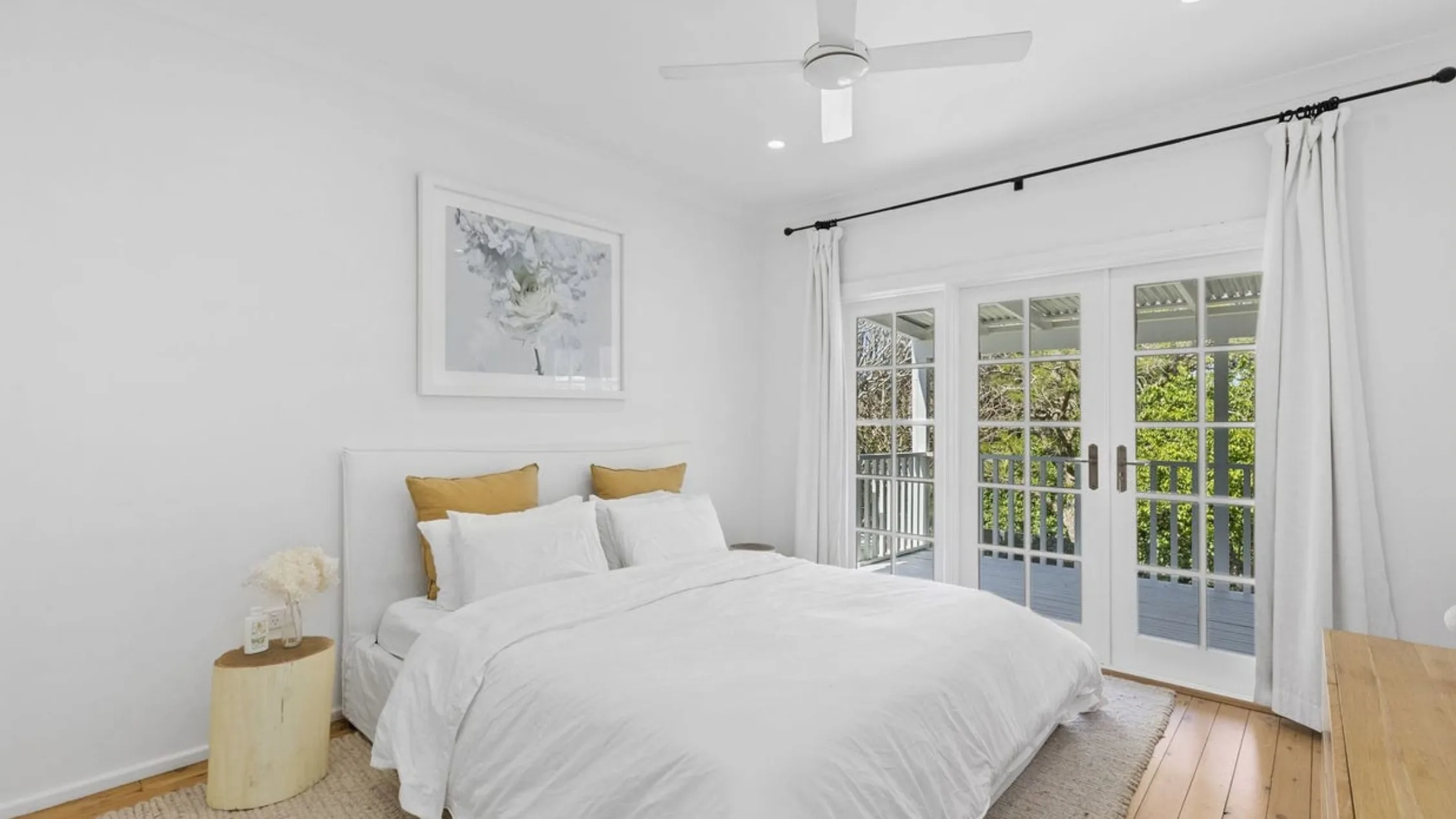 Eleanor Pendleton's White bedroom with a double bed, mustard pillows, wall art, glass doors, and a ceiling fan, leading to a balcony.