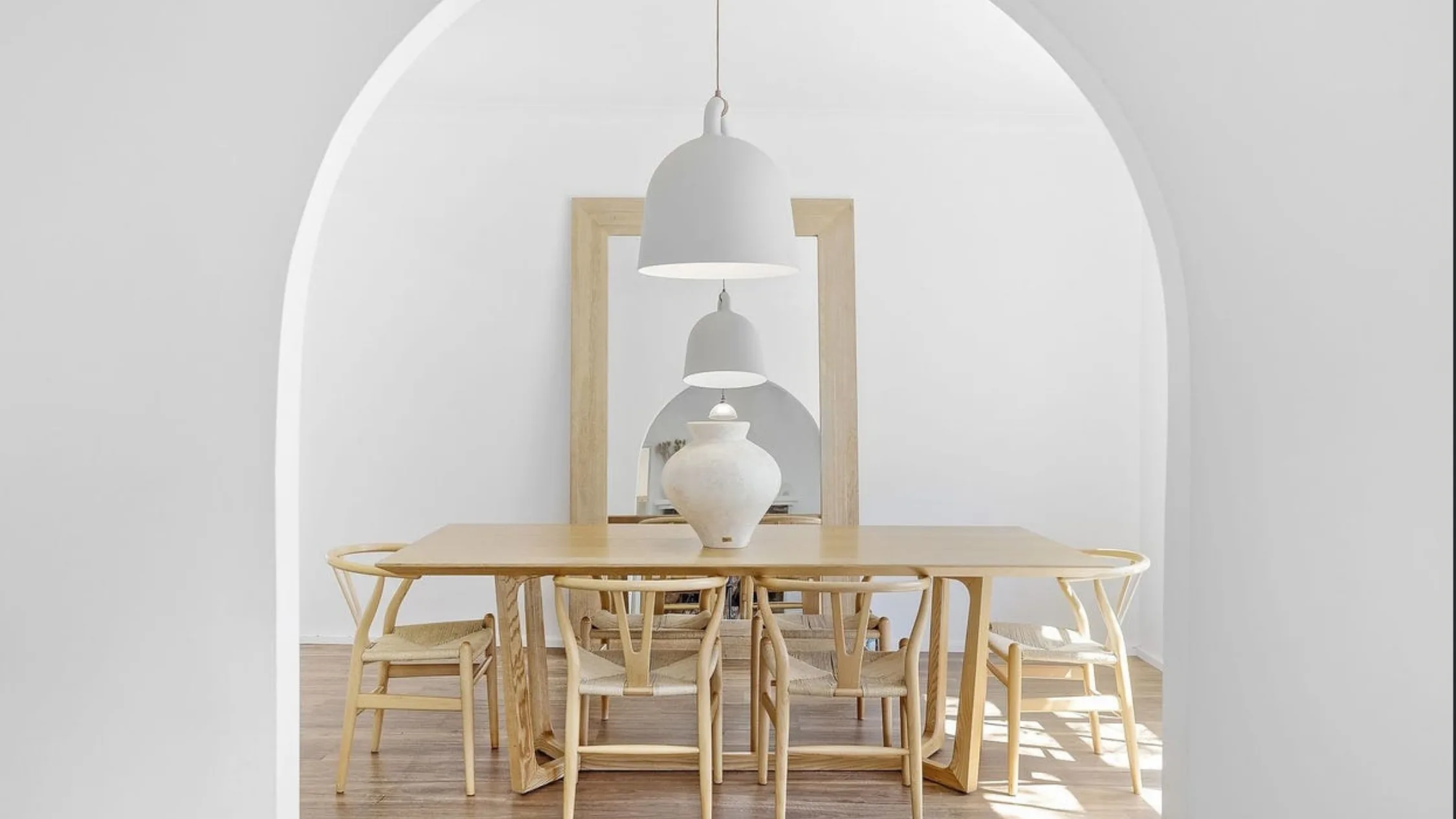 Eleanor Pendleton's Minimalist dining room with wooden table, chairs, large white pendant lamp, and vase, framed by an archway.