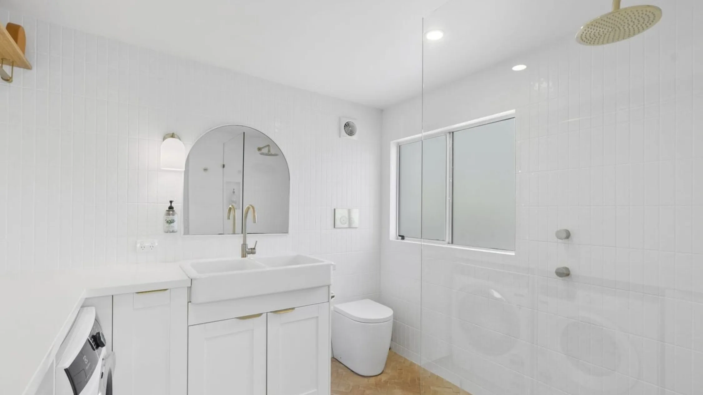 Eleanor Pendleton's Modern white bathroom with a sink, gold faucet, round mirror, washing machine, shower, and frosted window.