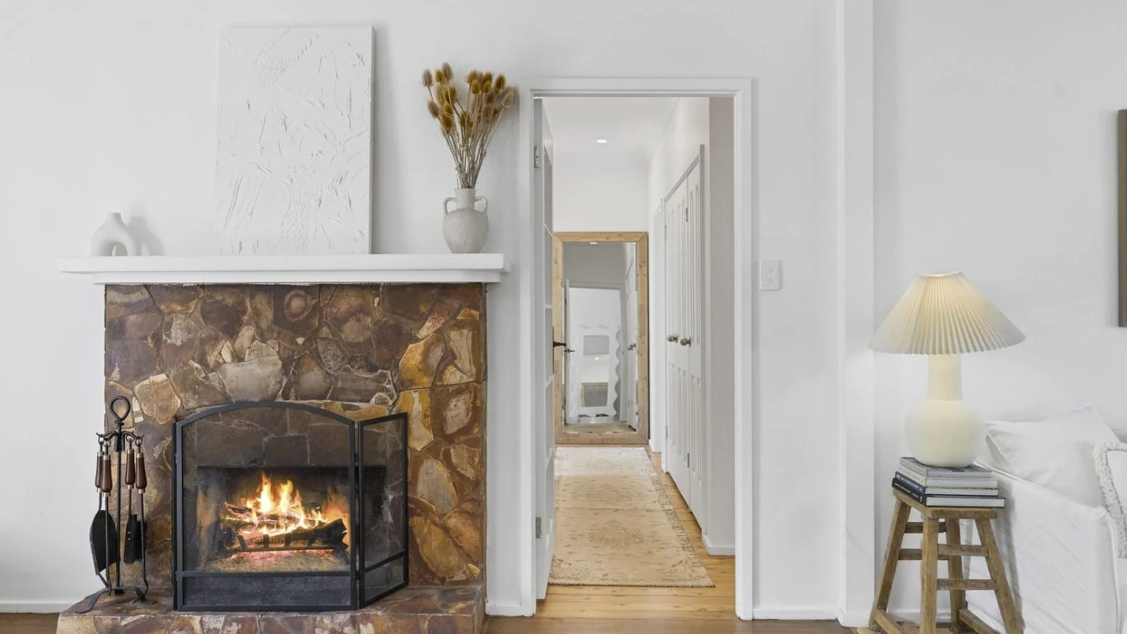 Eleanor Pendleton's Fireplace with stone surround in a living room, white walls, and a glowing lamp on a wooden stool beside a couch.