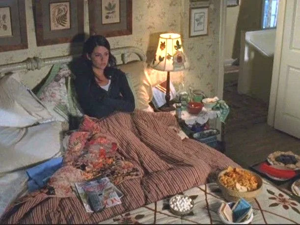 A still from the TV show Gilmore Girls. Lorelai Gilmore lies in her bed. She has bowls of snacks on her quilted bedspread, plus a magazine. Her bedside table is cluttered with food. Three botanical prints are framed above her metal bedhead.