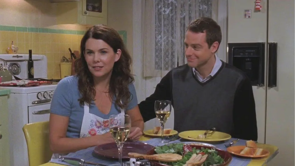 A still from the TV show Gilmore Girls. Lorelai and Christopher eat in Lorelai's kitchen. They are sitting at the dining table, which is yellow and rectangular. They sit on yellow dining chairs. Food is on the table in front of them, as well as wine glasses filled with white wine.