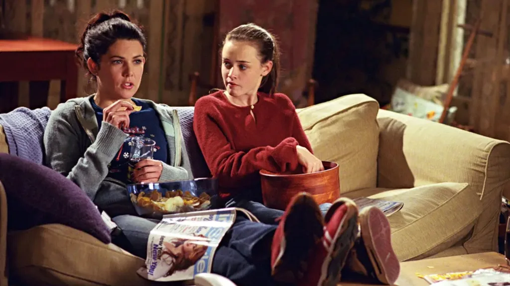 A still from the TV show Gilmore Girls. Lorelai and Rory sit on a cream coloured couch in their living room, eating chips and pretzels. An InStyle magazine sits on Lorelai's lap. Their feet are up on the wooden coffee table.