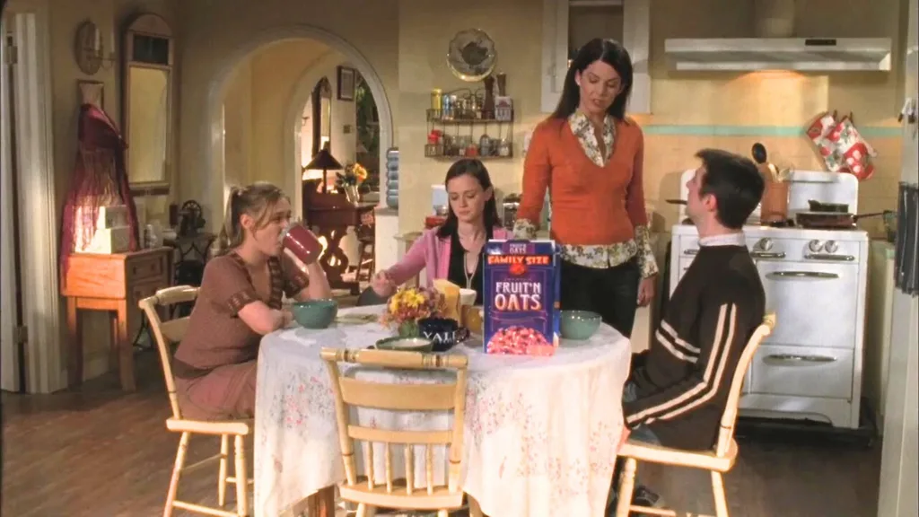 A still from the TV show Gilmore Girls. Rory, Kirk and Paris sit around the small circular dining table, positioned in the middle of the kitchen. The walls are yellow with a vintage white oven and cooker against one wall. Lorelai stands next to the table