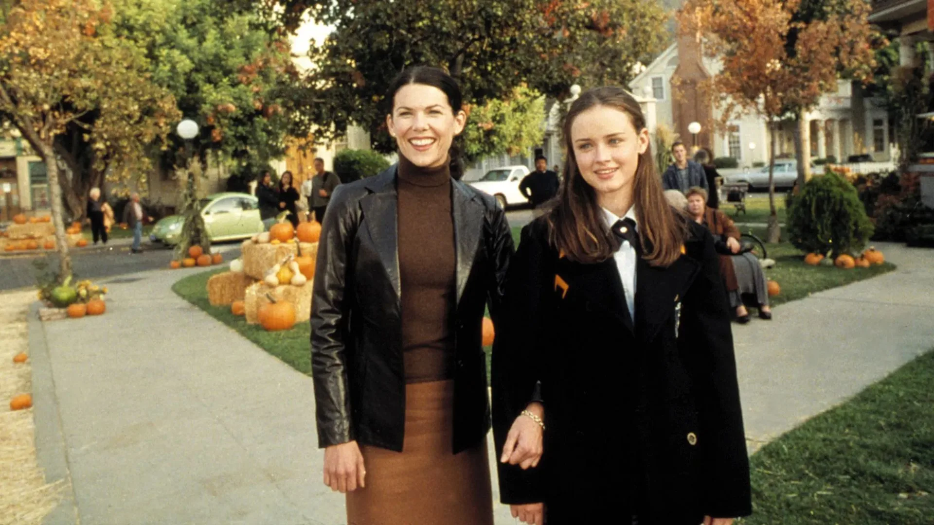 A still from the TV show Gilmore Girls. Rory and Lorelai walk through Stars Hollow, which has autumnal trees and is decorated with pumpkins.
