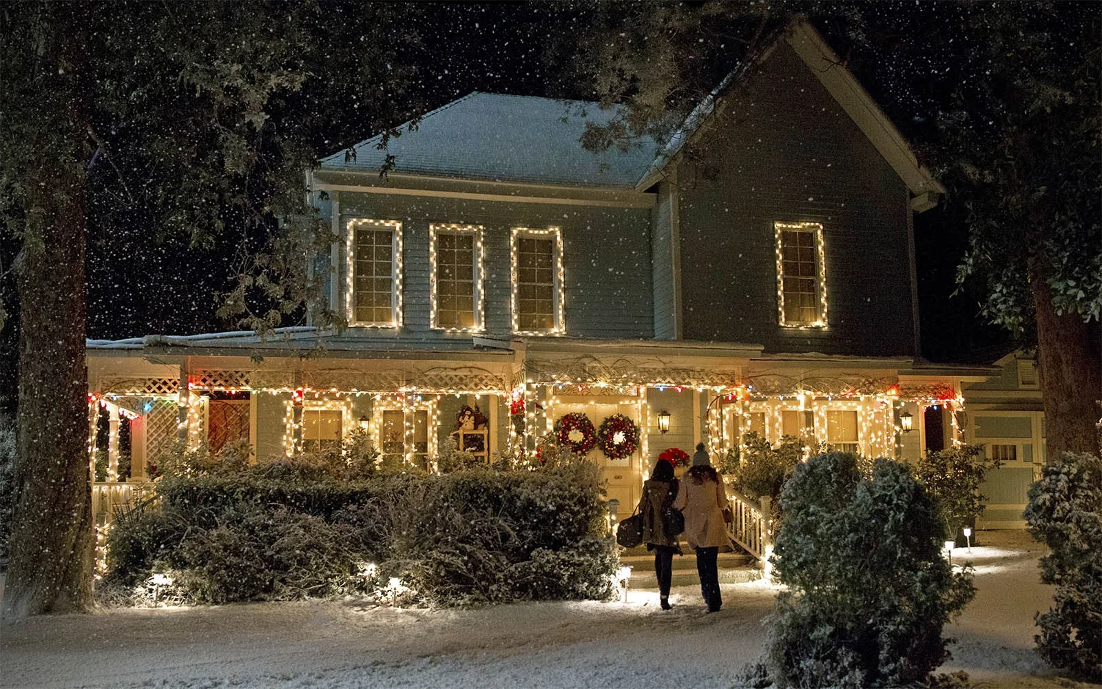 A still from the TV show Gilmore Girls: A Year In The Life. It shows the exterior of Lorelai and Rory's hosue which has been decorated with warm white Christmas lights, with two wreaths on the door. It is snowing.