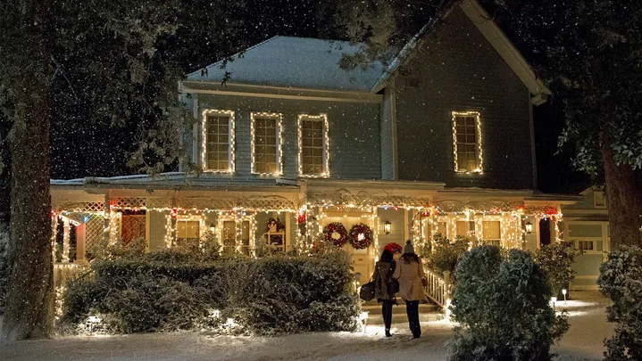A still from the TV show Gilmore Girls: A Year In The Life. It shows the exterior of Lorelai and Rory's hosue which has been decorated with warm white Christmas lights, with two wreaths on the door. It is snowing.
