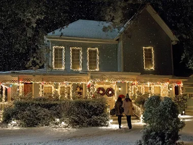A still from the TV show Gilmore Girls: A Year In The Life. It shows the exterior of Lorelai and Rory's hosue which has been decorated with warm white Christmas lights, with two wreaths on the door. It is snowing.