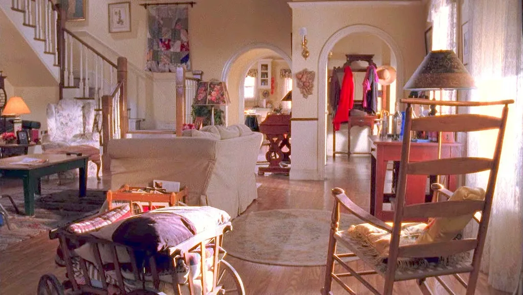 Cozy living room with rocking chair, staircase, and vintage decor, featuring a patchwork quilt hanging on the wall and a white couch. This is the living room of Lorelai and Rory in Gilmore Girls.