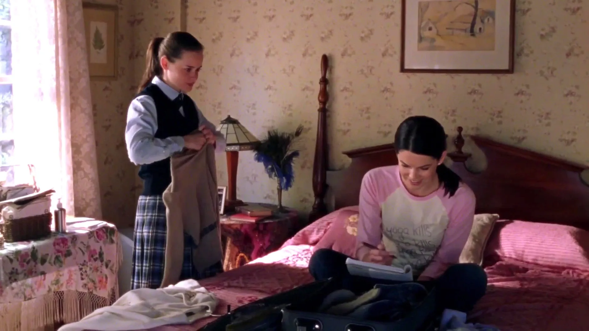 A still from the TV show Gilmore Girls, with Rory beside Lorelai's wooden bed. Lorelai sits on her bed, on pink sheets. A floral wallpaper covers the walls. A bedside table has a Tiffany lamp on it.