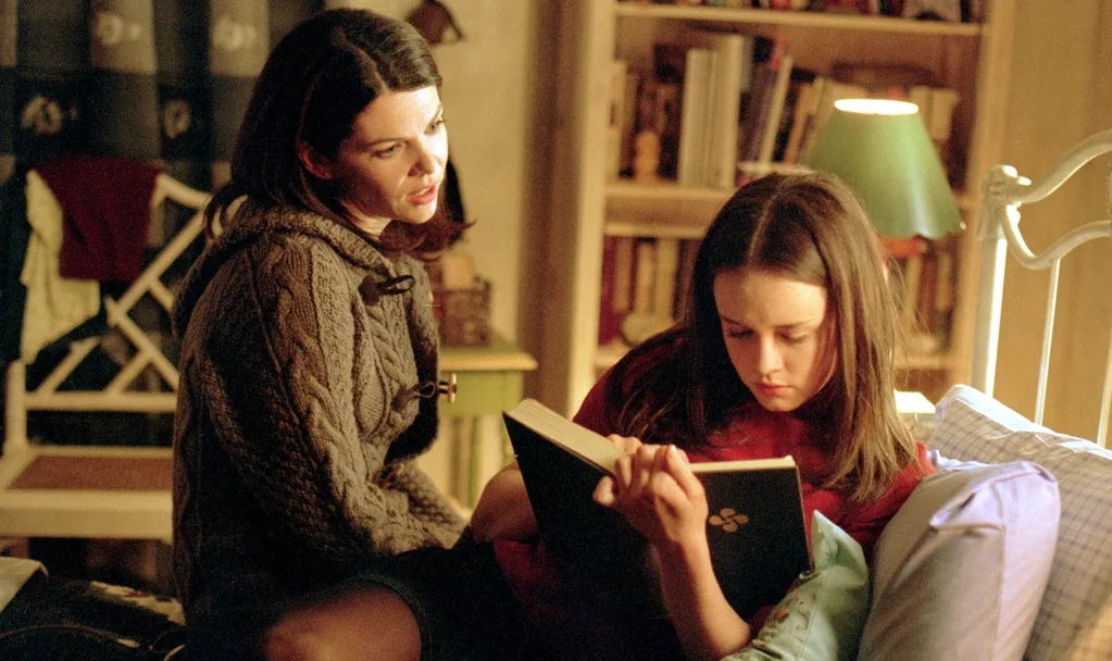 A still from the TV show Gilmore Girls. Rory is reading on her bed, Lorelai sits beside her. The bedframe is white and a bookcase filled with books is in the background, as is a white chair.