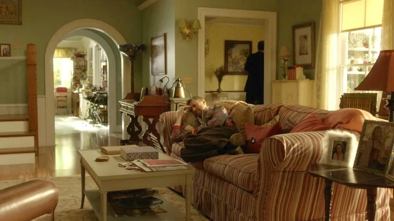 A still from the TV show Gilmore Girls: A Year In The Life. Character Kirk lays on a striped couch. A white coffee table is covered in books and sheets of paper. The walls are sage, with white arched doorways. There is a lot of vintage decor.
