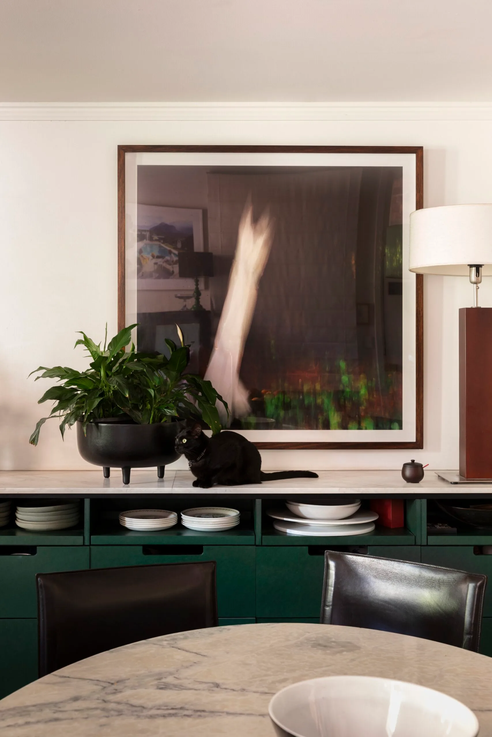 A kitchen and dining area with white benchtop running the length of the white wall and green cabinets underneath. A black cat sits on the benchtop, beside a potted plant. A round marble table with black dining chairs is positioned in front.