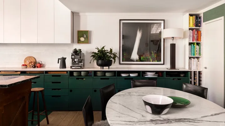 A kitchen with white countertops, green cabinets underneath the counter, white cabinets above the counter and a white tiled splashback. Coffee machine, plants and a lamp are on the countertop, while art is on the wall. A round marble table is positioned in front.