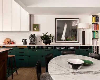 A kitchen with white countertops, green cabinets underneath the counter, white cabinets above the counter and a white tiled splashback. Coffee machine, plants and a lamp are on the countertop, while art is on the wall. A round marble table is positioned in front.