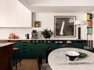 A kitchen with white countertops, green cabinets underneath the counter, white cabinets above the counter and a white tiled splashback. Coffee machine, plants and a lamp are on the countertop, while art is on the wall. A round marble table is positioned in front.