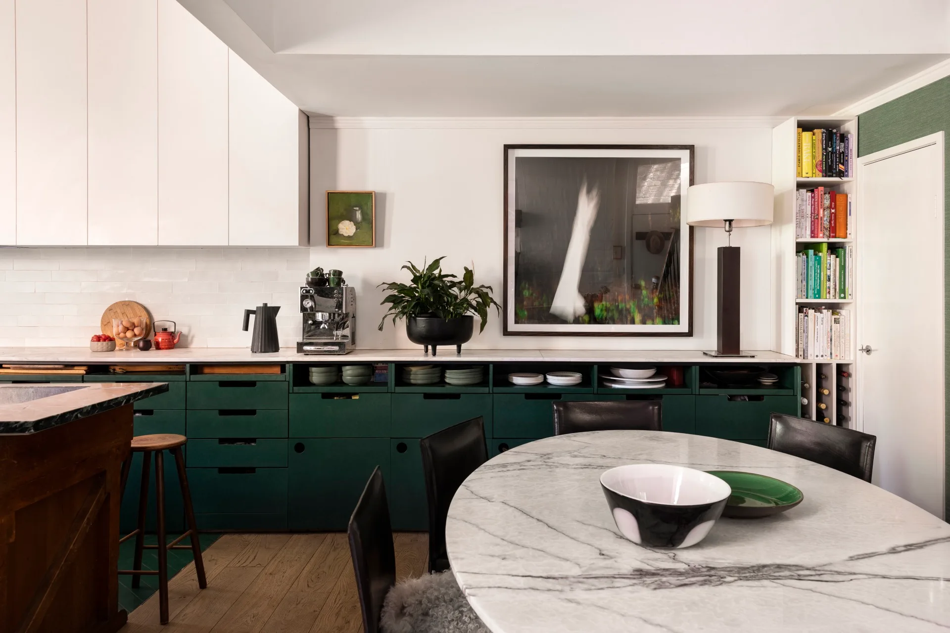 A kitchen with white countertops, green cabinets underneath the counter, white cabinets above the counter and a white tiled splashback. Coffee machine, plants and a lamp are on the countertop, while art is on the wall. A round marble table is positioned in front.