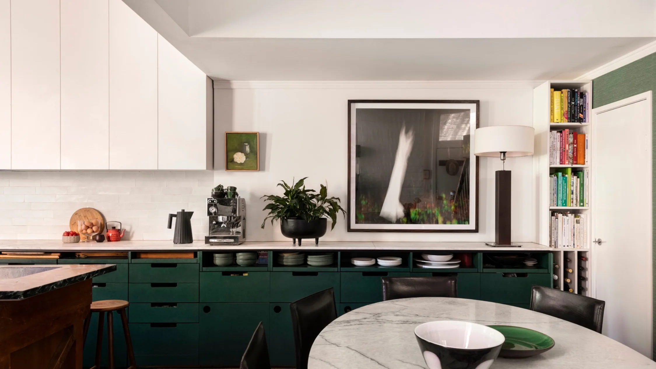 A kitchen with white countertops, green cabinets underneath the counter, white cabinets above the counter and a white tiled splashback. Coffee machine, plants and a lamp are on the countertop, while art is on the wall. A round marble table is positioned in front.