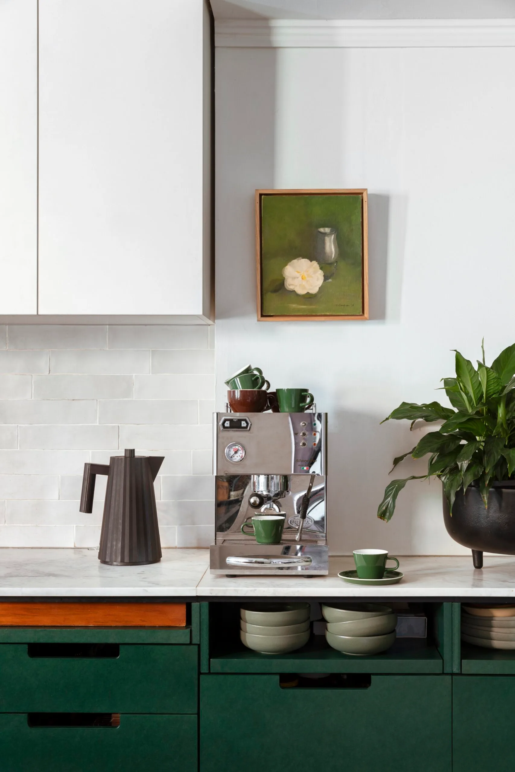 A kitchen with white countertops, green cabinets underneath the counter, white cabinets above the counter and a white tiled splashback. Coffee machine, plants and a jug are on the countertop, while art is on the wall.