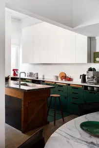 A u-shaped kitchen. Along one wall runs white benchtops, a white tiled splashback and white cabinets above the splashback. On the other side, a repurposed railway sleeper functions as a kitchen island, with a sink inside it. The benchtop of this is green marble. This complements the under-bench cabinetry, which is green.