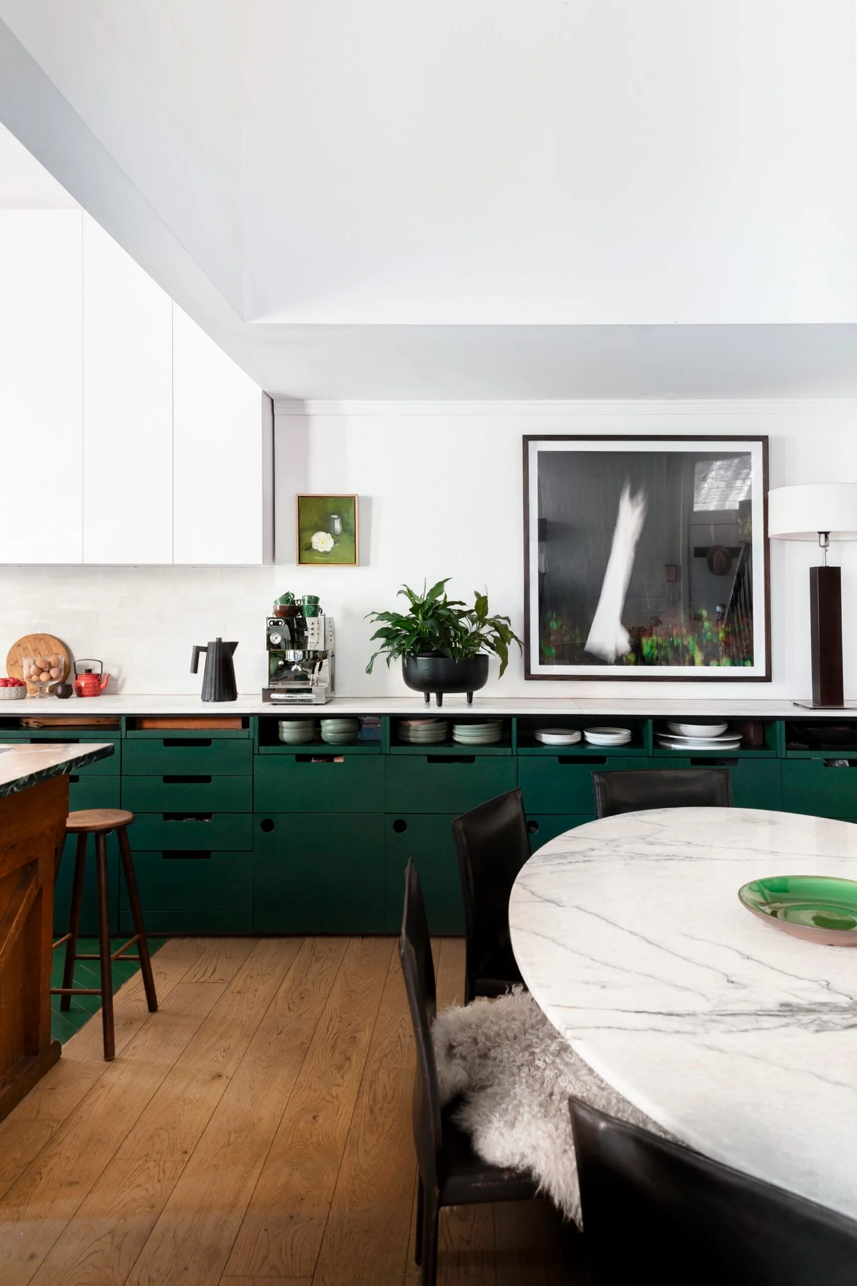 A kitchen with white countertops, green cabinets underneath the counter, white cabinets above the counter and a white tiled splashback. Coffee machine, plants and a lamp are on the countertop, while art is on the wall. A round marble table is positioned in front.