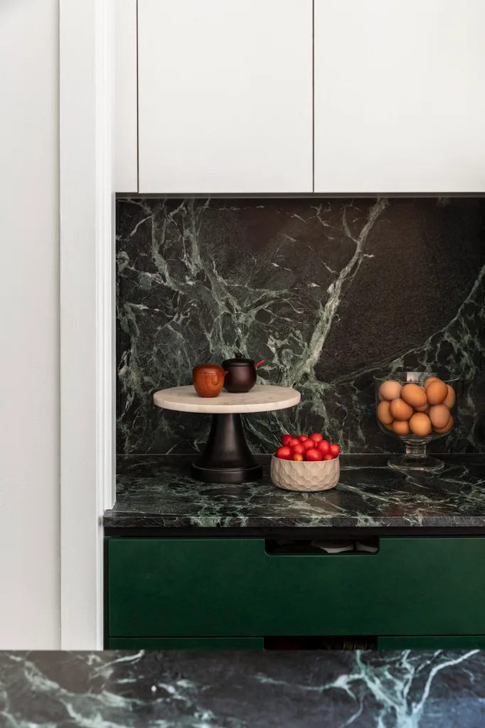 A close up photo of a section of a kitchen, with green marble benchtops and matching splashback. Green cabinetry is underneath the benchtop while white cabients are above the splashback. A cake stand and two bowls are positioned on the benchtop, filled with fruits, eggs and little jars.