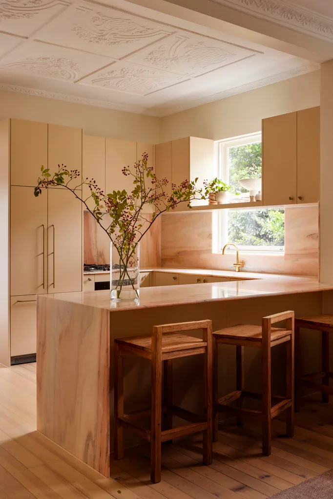 A kitchen in a heritage building that has a warm palette. The u-shaped kitchen has honey timber floors and bar stools, with warm cream toned joinery. Marble splashback and benchtop with a waterfall edge has a pink, peach and cream tone.