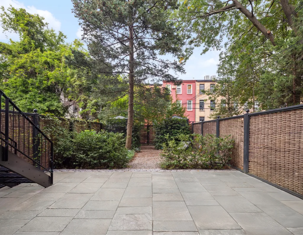 Paved patio with metal staircase, surrounded by trees and wicker fence, with colorful buildings in background.