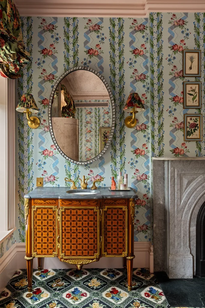 Vintage-style bathroom with floral wallpaper, ornate wooden vanity, oval mirror, patterned carpet, and twin brass wall sconces with pleated lampshades.