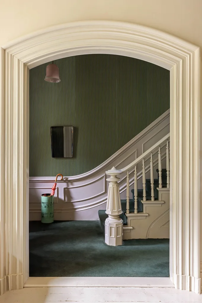 A green-carpeted hallway with stairs, ornate white banister, striped green wallpaper above white wainscoting, mirror, pink umbrella in holder, and vintage lampshade.