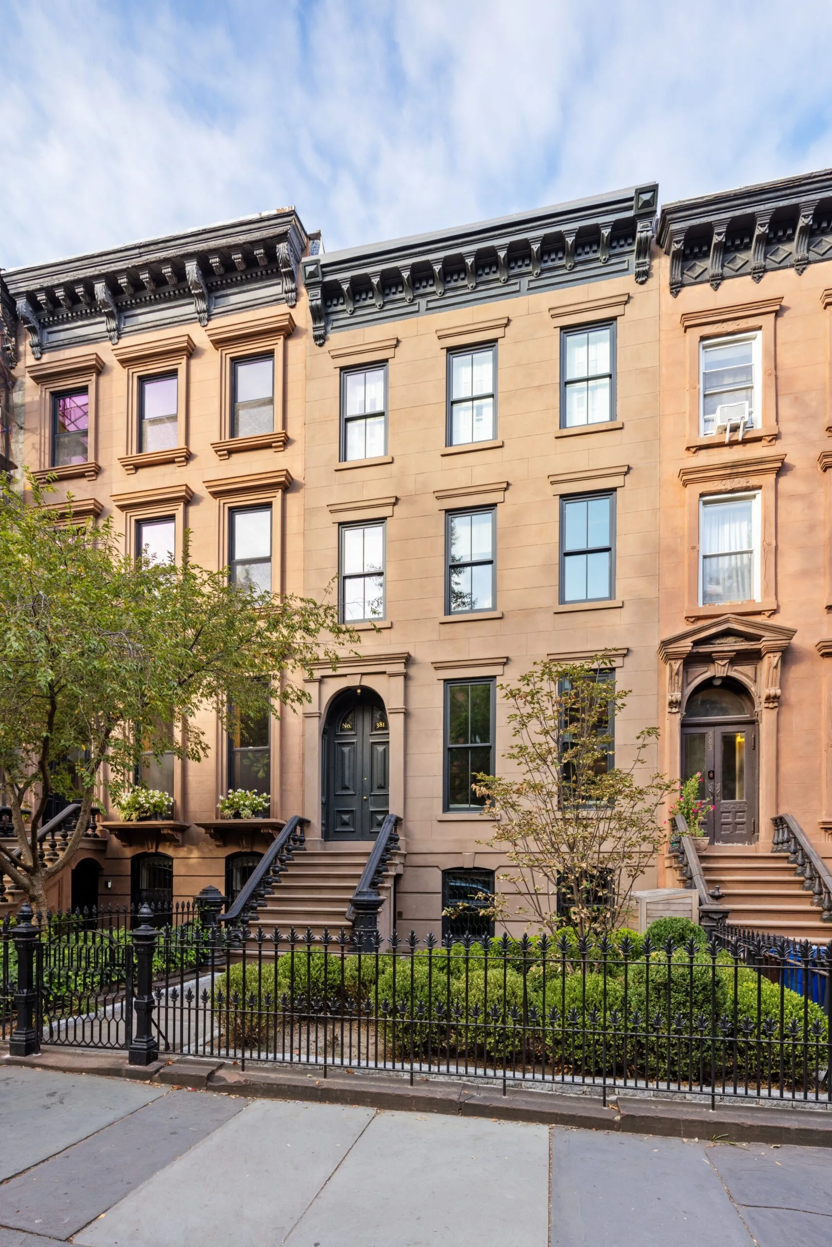 Brownstone building with black doors and steps, iron railing, and small trees in front on a clear day. This is the exterior of Lily Allen and David Harbour's house.