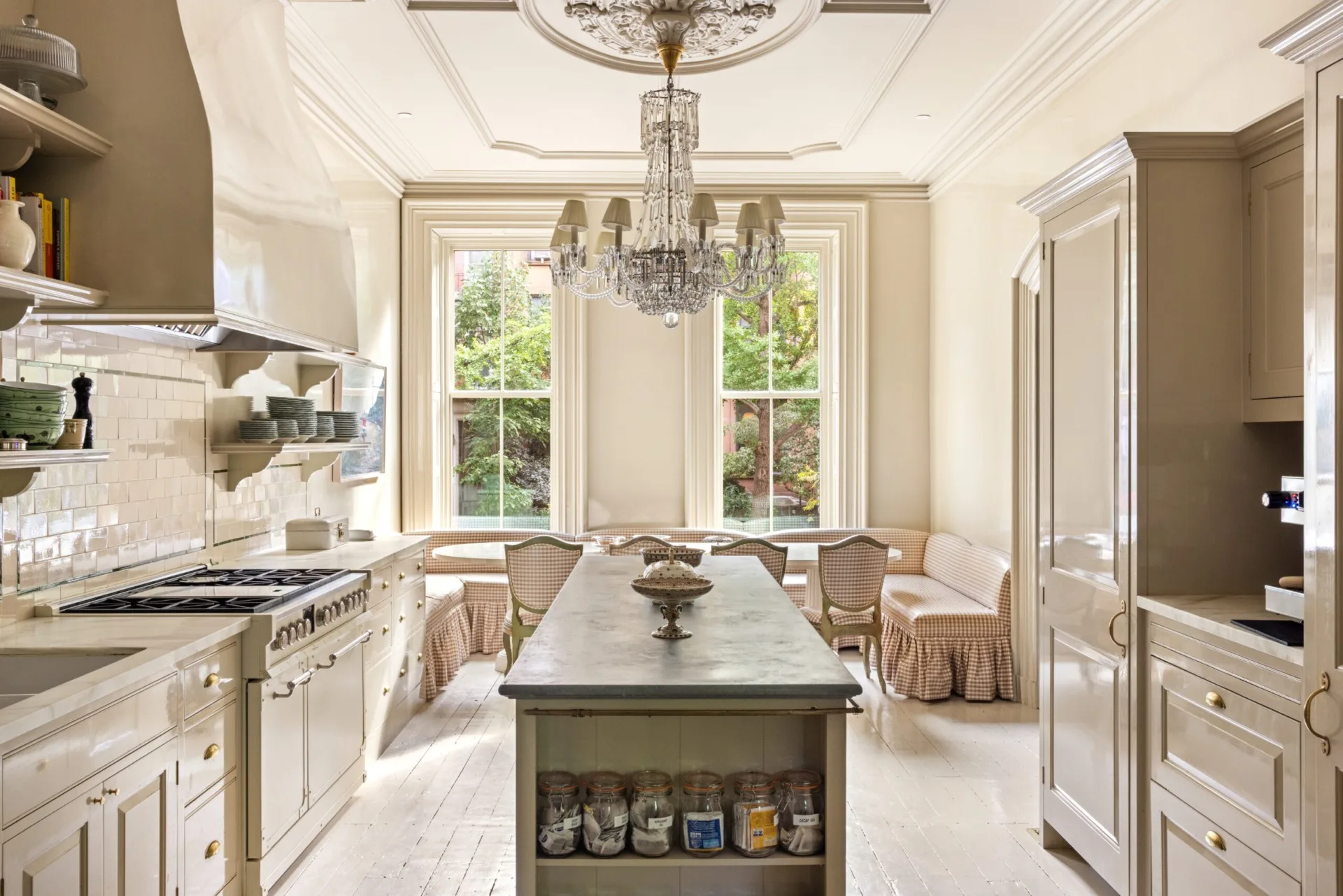 Luxurious kitchen with a chandelier, central island, large windows, and a cozy dining nook with upholstered banquette seating. It features Plain English cabinetry, which Lily Allen wanted.