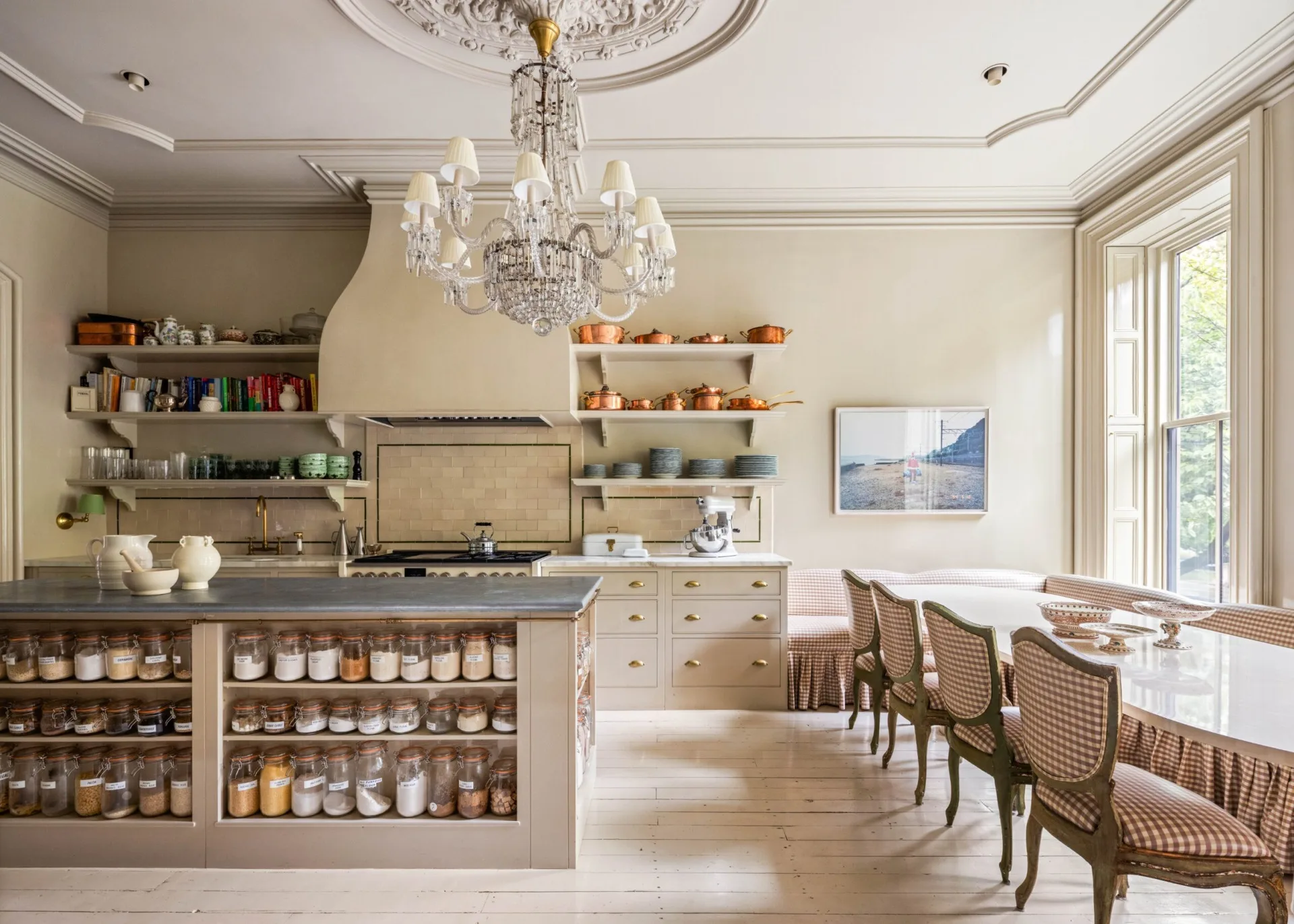 Luxurious kitchen with chandelier, open shelves, glass jars, and vintage dining set, against large windows and light decor. The right side of the kitchen has a built-in banquette with a long, oval table.