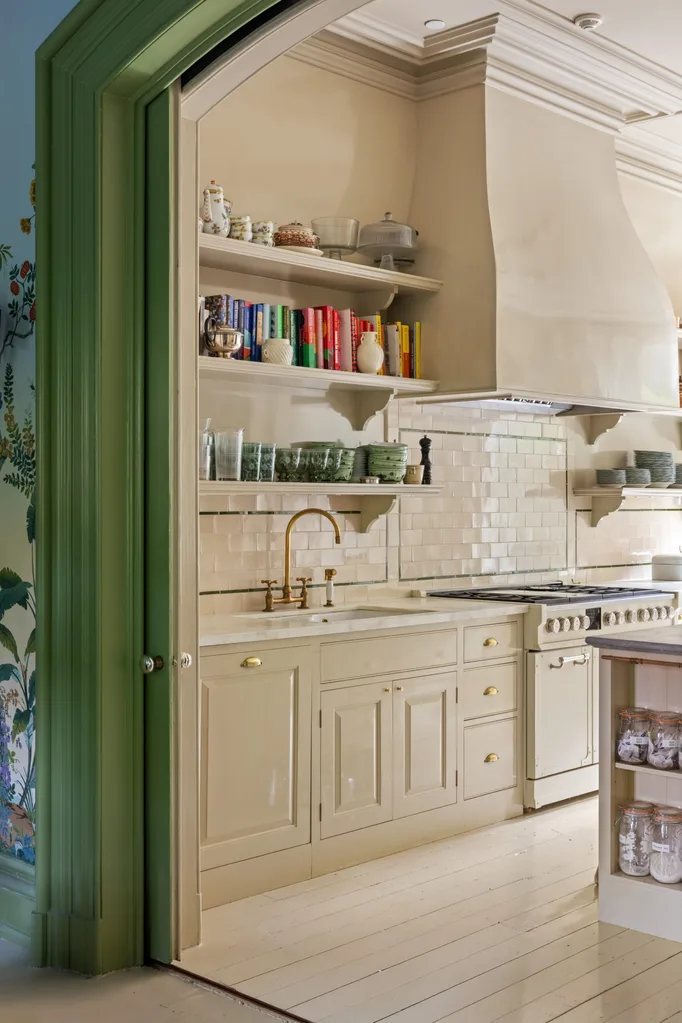 A cozy kitchen with cream cabinets, brass faucet, open shelves with books and dishes, tiled splashback, and green door frame.