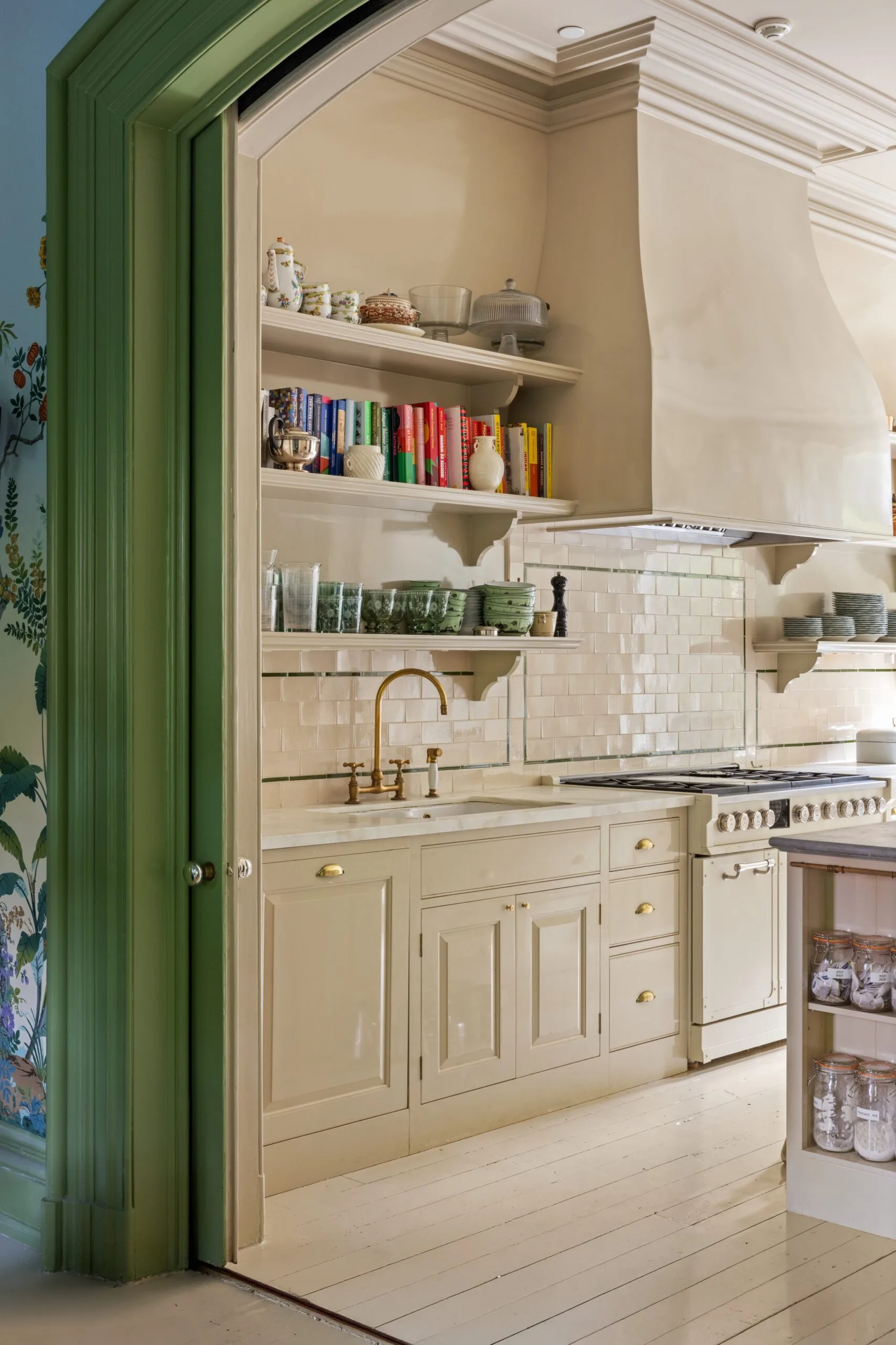 A cozy kitchen with cream cabinets, brass faucet, open shelves with books and dishes, tiled splashback, and green door frame.